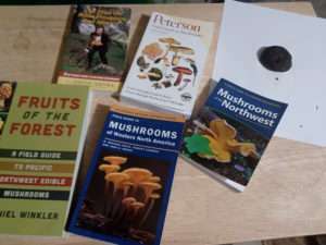 An assortment of field guides to mushrooms relevant to the Pacific Northwest sitting on a wooden table, plus a piece of white paper with a mushroom cap sitting on it, for article on how to identify mushrooms