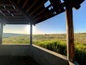 A view from underneath the roof of a wooden gazebo overlooking a wide grassy plain in early evening with the sun low in the blue sky.